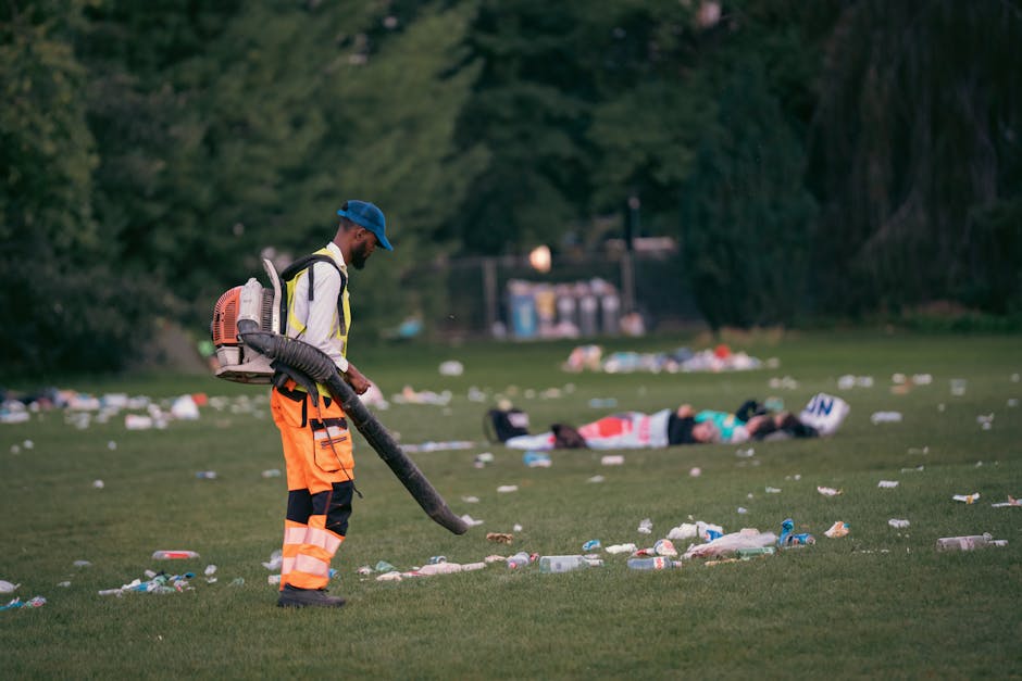 A male waste management worker wearing high-visibility orange trousers, a white jacket with reflective stripes, and a blue cap is using a leaf blower or similar equipment to clear up scattered litter across a grassy park area. The worker is positioned to the left of the image, focused on the task of collecting rubbish such as plastic bottles, paper waste, and other debris, which are spread unevenly across the open space. In the background, there are a few people lying on the grass, with a variety of trash around them, suggesting recent littering or a site of an event. Mature trees and bushes border the park, and a small utility area with containers can be seen at a distance. The scene appears to be late in the day or early evening, with diffuse natural lighting providing a clear view of the litter cleanup process. This image highlights the importance of professional rubbish removal services, like those offered by Rubbish Removal Belsize Park, for maintaining clean outdoor environments through on-site clearance and private waste handling solutions.