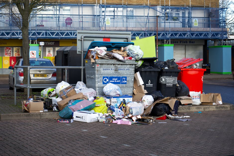 An outdoor scene showing a collection of overflowing rubbish and discarded waste placed on the pavement in front of a building with a blue scaffolding structure. The waste includes various black, black-and-white, and red plastic rubbish bags, some ripped open revealing cardboard boxes, paper, and plastic packaging inside. There are large grey and black wheelie bins, with one lid open and some waste spilling out, alongside flattened cardboard boxes and small discarded items scattered across the ground. A parked silver hatchback car is nearby, partly obscured by a metal railing, and the background features shop fronts with closed shutters and signage. The environment is an urban setting indicating an incident of inadequately managed rubbish collection or on-site waste disposal, consistent with independent waste handling services such as those provided by Rubbish Removal Belsize Park. The natural lighting suggests daytime with overcast sky, and the scene emphasizes the necessity for professional rubbish removal services to handle such waste accumulation effectively and safely.