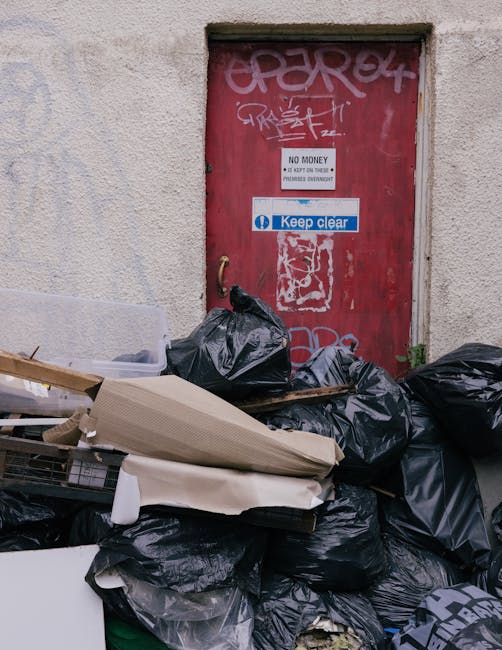 An outdoor scene showing a collection of overflowing rubbish and discarded waste placed on the pavement in front of a building with a blue scaffolding structure. The waste includes various black, black-and-white, and red plastic rubbish bags, some ripped open revealing cardboard boxes, paper, and plastic packaging inside. There are large grey and black wheelie bins, with one lid open and some waste spilling out, alongside flattened cardboard boxes and small discarded items scattered across the ground. A parked silver hatchback car is nearby, partly obscured by a metal railing, and the background features shop fronts with closed shutters and signage. The environment is an urban setting indicating an incident of inadequately managed rubbish collection or on-site waste disposal, consistent with independent waste handling services such as those provided by Rubbish Removal Belsize Park. The natural lighting suggests daytime with overcast sky, and the scene emphasizes the necessity for professional rubbish removal services to handle such waste accumulation effectively and safely.