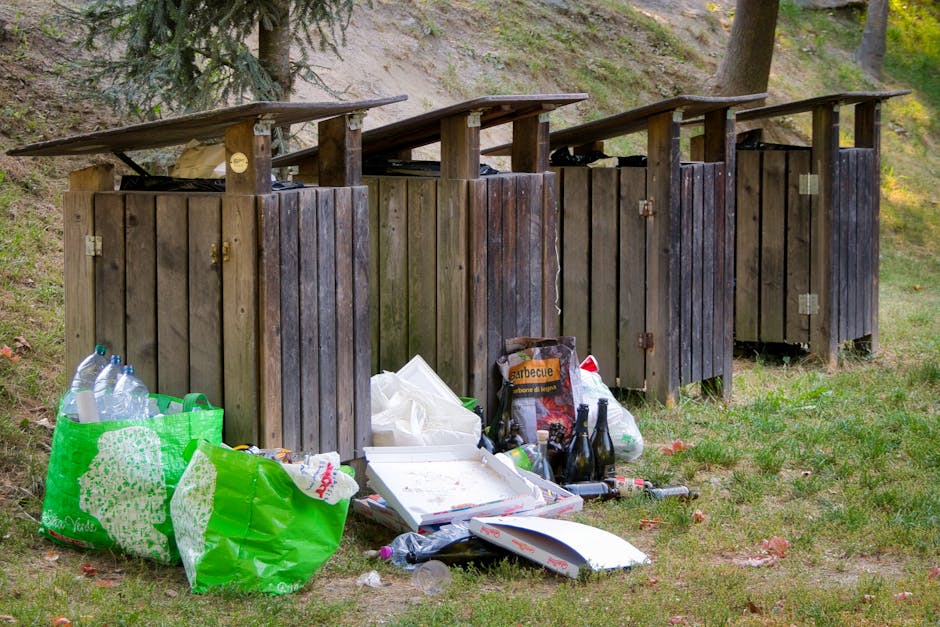 Four wooden rubbish bins with sloped, weathered dark brown and light brown panel doors are lined up on a grassy area, situated in front of a sloped, grassy hillside with trees and bushes in the background. The bins are partially open at the top, with gaps and visible hinges on the doors, indicating they are meant for waste collection. In front of the bins, various waste items are scattered on the grass, including two large green plastic bags filled with empty plastic bottles, a white cardboard pizza box with some food residue, a black plastic bag, a few empty glass bottles (including one with a red label), and other miscellaneous rubbish such as plastic and paper debris. The environment appears to be an outdoor setting, possibly a park or a garden area, with natural daylight illuminating the scene. The scene illustrates an example of private waste disposal through an independent rubbish clearance service, as provided by Rubbish Removal Belsize Park, highlighting the condition of unmanaged waste near outdoor rubbish bins.
