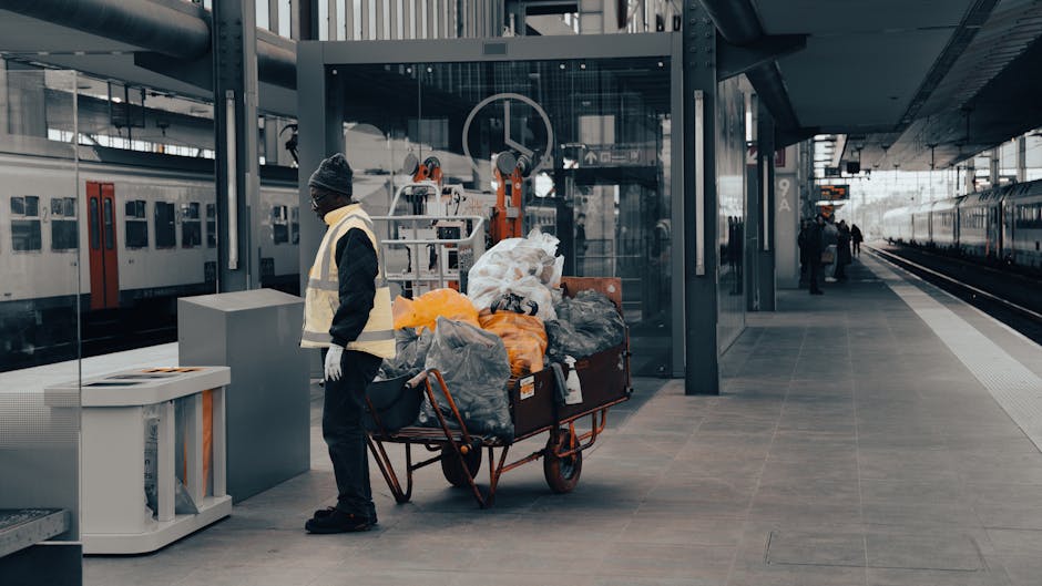 A man wearing a dark jacket, grey beanie, and white gloves stands next to a large red trolley loaded with various bags and loose rubbish, including black, grey, and orange refuse sacks, as well as plastic and paper debris. The trolley is positioned on a tiled platform at a modern train station, with a glass-enclosed lift and a train with closed doors visible behind him. To the right, the platform continues towards a distant train track, with a few other passengers in the background. The scene is lit by natural daylight, highlighting the urban environment and clean, structured architectural elements. The presence of the trolley and rubbish bags suggests the man may be involved in waste collection or rubbish removal, consistent with services offered by Rubbish Removal Belsize Park for alternative or private waste handling near Belsize Park Station.