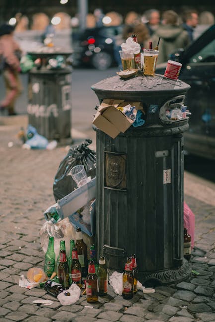 A black, cast-iron rubbish bin with a rounded, domed lid is positioned on a cobbled street. The lid is cluttered with discarded paper cups, plastic cups with leftover drinks, and crumpled food packaging, some of which are leaning on the bin's surface. Surrounding the base of the bin, there are multiple empty glass beer bottles, some upright and others lying on the cobblestones, along with crumpled paper, plastic wrappers, and other miscellaneous litter. A black trash bag is partially visible next to the bin, with some overflow of waste spilling onto the street. In the background, blurred pedestrians can be seen walking past, along with parked cars, indicating a busy urban environment. The scene appears to be during daytime with natural lighting, emphasizing the littered state of this outdoor public space, highlighting the need for waste collection services such as those provided by Rubbish Removal Belsize Park, which specialize in managing waste from busy street areas and private disposal needs.