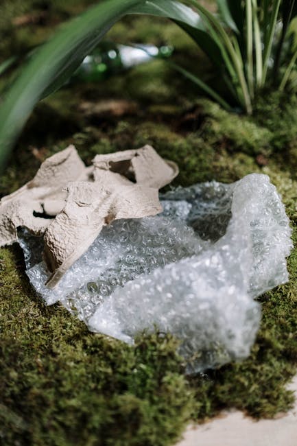 A close-up photograph of a broken disposable coffee cup made from light brown, textured cardboard resting on a bed of green moss. Surrounding the cup fragments are clear, crumpled pieces of plastic bubble wrap, partially stretched and layered on the mossy surface. In the background, there are lush green leaves and foliage, suggesting the scene is outdoors in a garden or natural setting. The lighting appears natural, highlighting the textures of the moss, cardboard, and plastic, emphasizing the contrast between organic and synthetic materials, which relates to waste disposal and rubbish removal services such as those provided by Rubbish Removal Belsize Park. The scene reflects environmentally conscious waste handling with an emphasis on non-recyclable packaging and packaging debris often seen in private disposal or on-site clearance contexts.