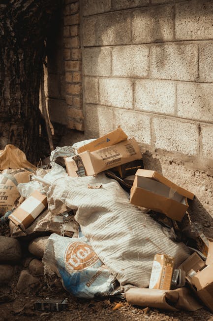 A black, cast-iron rubbish bin with a rounded, domed lid is positioned on a cobbled street. The lid is cluttered with discarded paper cups, plastic cups with leftover drinks, and crumpled food packaging, some of which are leaning on the bin's surface. Surrounding the base of the bin, there are multiple empty glass beer bottles, some upright and others lying on the cobblestones, along with crumpled paper, plastic wrappers, and other miscellaneous litter. A black trash bag is partially visible next to the bin, with some overflow of waste spilling onto the street. In the background, blurred pedestrians can be seen walking past, along with parked cars, indicating a busy urban environment. The scene appears to be during daytime with natural lighting, emphasizing the littered state of this outdoor public space, highlighting the need for waste collection services such as those provided by Rubbish Removal Belsize Park, which specialize in managing waste from busy street areas and private disposal needs.