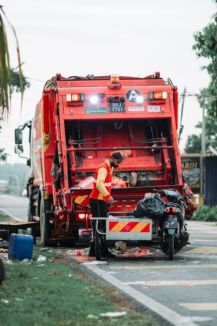 A worker wearing a yellow high-visibility vest and black trousers is standing behind the rear of a large red rubbish collection truck, which is parked on the side of a street. The worker appears to be handling waste or equipment on the open rear-loading mechanism, which is partially raised, exposing the interior of the bin area. The truck has various safety and identification labels, including a white circular symbol and the registration number 'BRJ 7751.' Nearby, there is a small black motorcycle parked close to the truck, with a plastic bag placed on its seat or rear cargo area. In the foreground, a metal waste container or bin with a red and white striped reflector panel is positioned on the street edge. The surrounding environment includes a grassy verge with a small blue water container or recycling bin, and a background featuring a sidewalk, some trees, and a building or roadside advertisement board. Overcast daylight creates a diffuse lighting atmosphere, emphasizing the details of the vehicle and the worker engaged in waste collection, reflecting an image related to private waste handling or rubbish removal services by Rubbish Removal Belsize Park.